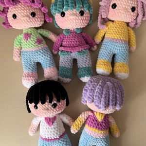 K&ouml;nnte beinhalten: Sechs handgefertigte H&auml;kelpuppen mit verschiedenen Haarfarben und Outfits. Jede Puppe hat ein rundes Gesicht, schwarze Augen und rosige Wangen. Die Puppen tragen bunte Oberteile und Hosen mit Details wie Schleifen und Sternen. Die Puppen sind aus weichem, strukturiertem Garn gefertigt.