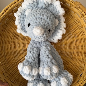 Peut inclure: Un jouet dinosaure Tricératops fait à la main, crocheté en bleu clair et blanc. Le dinosaure a une texture duveteuse, des yeux noirs et des accents blancs sur ses cornes et ses pieds. Il est posé dans un panier en osier.