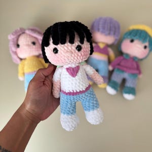 K&ouml;nnte beinhalten: Eine Sammlung handgefertigter H&auml;kelpuppen mit verschiedenen Haarfarben und Outfits. Die zentrale Puppe hat schwarzes Haar, ein wei&szlig;es Hemd mit einem rosa Herz und blaue Hosen. Andere Puppen haben rosa, lila und t&uuml;rkisfarbenes Haar mit passender Kleidung.
