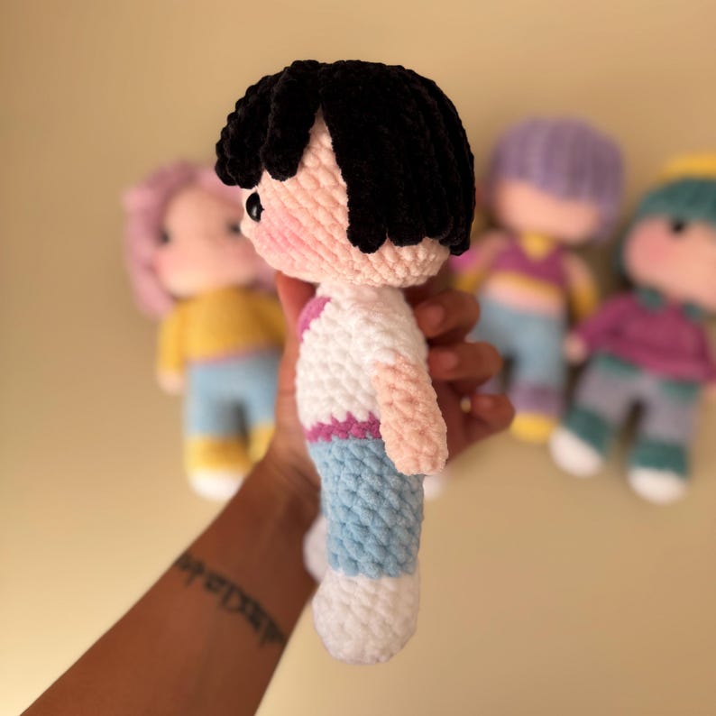 K&ouml;nnte beinhalten: Handgemachte H&auml;kelpuppe mit schwarzen Haaren, rosigen Wangen und einem wei&szlig;en Hemd mit rosa und blauen Akzenten. Die Puppe tr&auml;gt eine blaue Hose und wei&szlig;e Schuhe. Weitere Puppen sind im Hintergrund zu sehen.