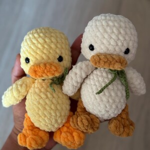 May include: Two handmade crochet ducklings. One is yellow with orange feet and beak, the other is white with orange feet and beak. Both have black eyes and green accents around their necks. These plush toys are soft and cuddly.