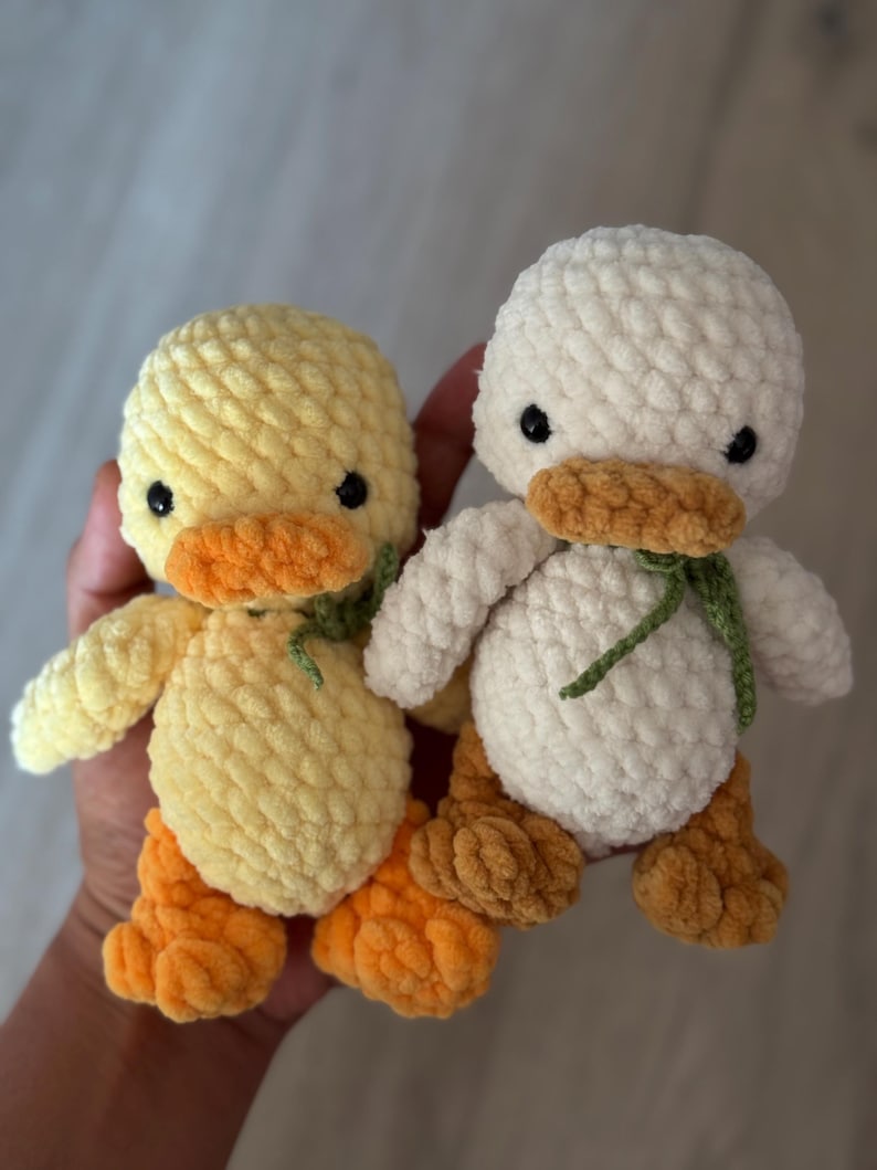 May include: Two handmade crochet ducklings. One is yellow with orange feet and beak, the other is white with orange feet and beak. Both have black eyes and green accents around their necks. These plush toys are soft and cuddly.
