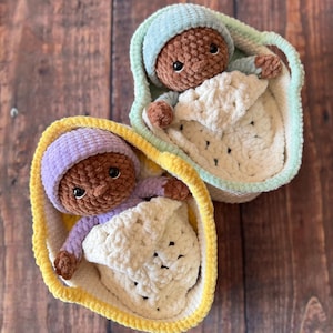 May include: Two handmade crochet baby dolls, each nestled in a cozy basket. One doll wears a purple hat and lavender outfit, the other a blue hat and light blue outfit. Both are wrapped in a cream-colored blanket, creating a soft and comforting scene.
