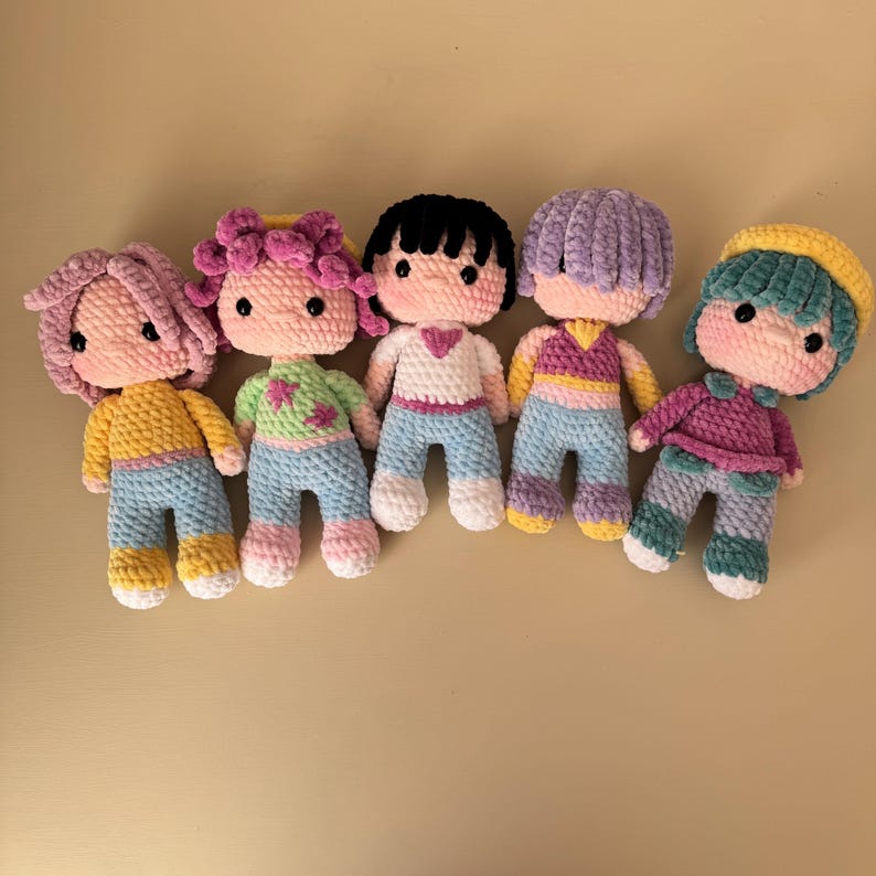 K&ouml;nnte beinhalten: F&uuml;nf handgefertigte H&auml;kelpuppen mit unterschiedlichen Haarfarben und Outfits. Jede Puppe hat ein einzigartiges Farbschema, darunter rosa, lila, schwarzes und t&uuml;rkisfarbenes Haar. Die Puppen tragen verschiedene Oberteile und Hosen mit Details wie Herzen und Streifen.