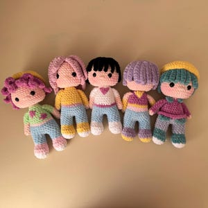 K&ouml;nnte beinhalten: F&uuml;nf handgefertigte H&auml;kelpuppen mit unterschiedlichen Haarfarben und Outfits. Jede Puppe hat ein einzigartiges Farbschema, darunter rosa, schwarze, lila und gelbe Haare sowie Outfits in Blau-, Gelb-, Gr&uuml;n- und Rosat&ouml;nen. Die Puppen sind in einer Reihe angeordnet.