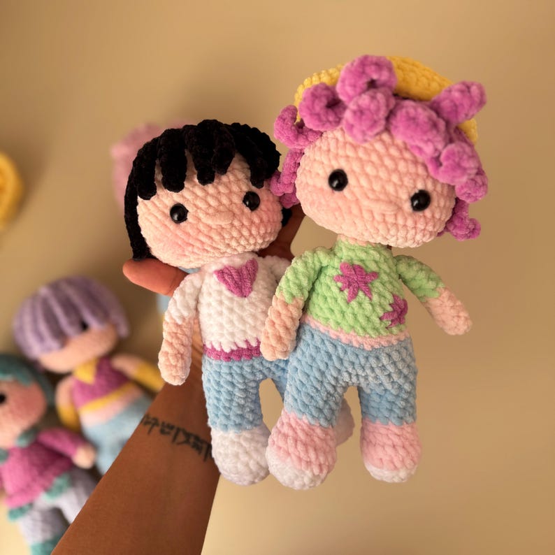 K&ouml;nnte beinhalten: Zwei handgefertigte H&auml;kelpuppen mit schwarzen und rosa Haaren, die wei&szlig;e Hemden, blaue Hosen und rosa Schuhe tragen. Die Puppen haben schwarze Knopfaugen und werden vor einem verschwommenen Hintergrund mit anderen &auml;hnlichen Puppen gehalten.