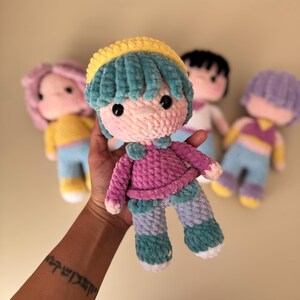 K&ouml;nnte beinhalten: Handgemachte H&auml;kelpuppe mit rosa Gesicht, t&uuml;rkisfarbenem Haar und gelbem Hut. Die Puppe tr&auml;gt einen rosa Pullover, t&uuml;rkisfarbene und blaue Hosen und hat schwarze Knopfaugen. Andere Puppen mit unterschiedlichen Haarfarben sind im Hintergrund.