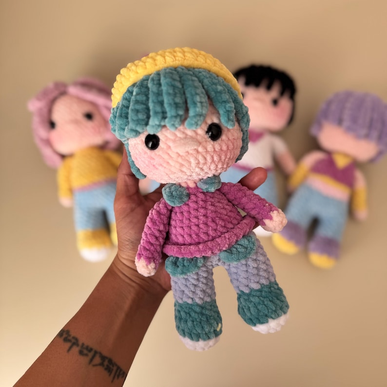 K&ouml;nnte beinhalten: Handgemachte H&auml;kelpuppe mit rosa Gesicht, t&uuml;rkisfarbenem Haar und gelbem Hut. Die Puppe tr&auml;gt einen rosa Pullover, t&uuml;rkisfarbene und blaue Hosen und hat schwarze Knopfaugen. Andere Puppen mit unterschiedlichen Haarfarben sind im Hintergrund.