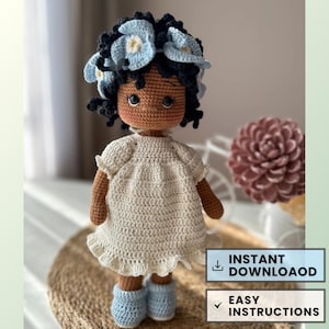 May include: Crochet doll pattern featuring a doll with brown skin, black curly hair, and a white dress. The doll is wearing a blue flower crown and blue booties. The pattern includes easy instructions and is available for instant download.