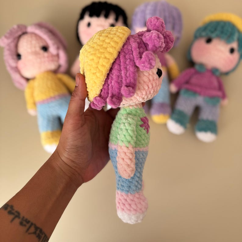 K&ouml;nnte beinhalten: Eine Sammlung handgefertigter H&auml;kelpuppen aus weichem Garn. Die Puppen haben farbenfrohe Outfits, darunter gelbe H&uuml;te, rosa und lila Haare und pastellfarbene Kleidung. Jede Puppe hat ein einzigartiges Design, ideal f&uuml;r Kinder.