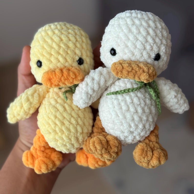 May include: Two plush duck toys, one yellow and one white, with orange beaks and feet. Each duck has black eyes and a green ribbon around its neck. The ducks are handmade and have a soft, textured appearance.