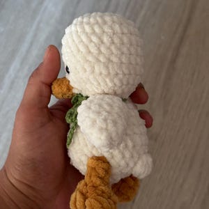 May include: A handmade, plush white duck with orange feet and beak, and a green scarf. The soft, textured duck is a cuddly toy, perfect for a child's room or as a decorative item. The duck is approximately 6 inches tall.