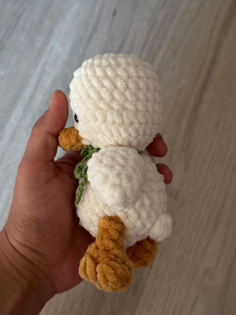 May include: A handmade, plush white duck with orange feet and beak, and a green scarf. The soft, textured duck is a cuddly toy, perfect for a child's room or as a decorative item. The duck is approximately 6 inches tall.