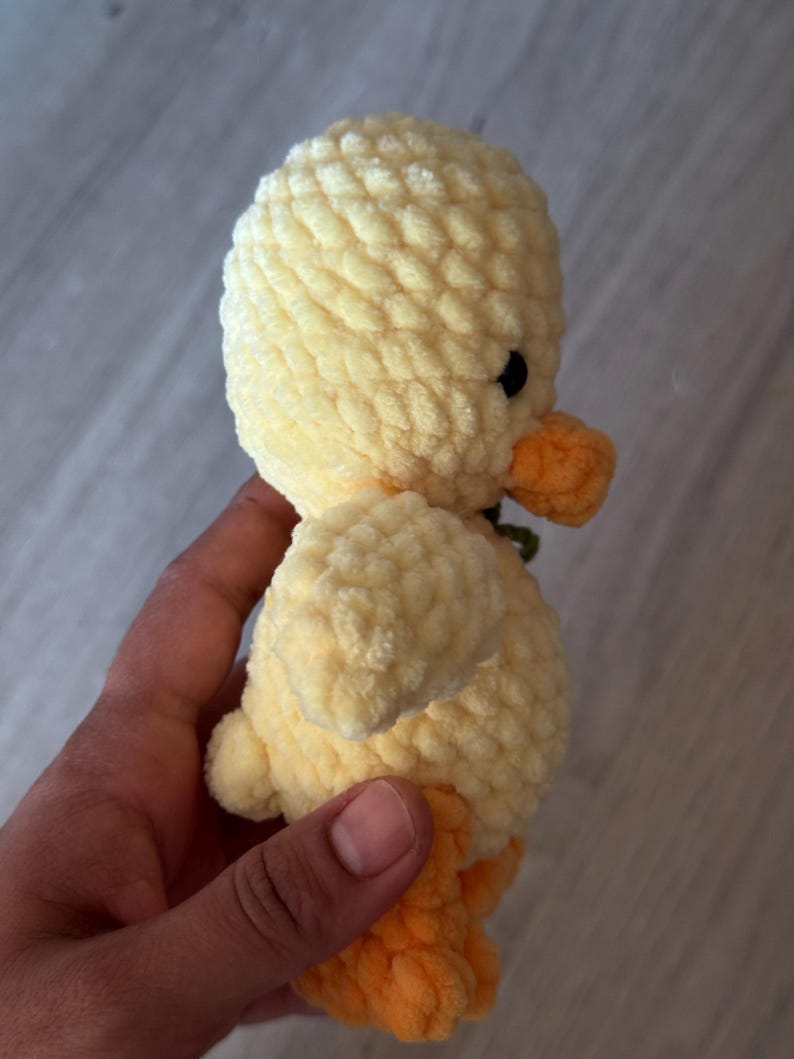 May include: A handmade, plush yellow duckling with an orange beak and feet. The duckling has small black eyes and a textured, soft appearance. The toy is likely crocheted or knitted, and is a cute, cuddly item.