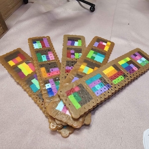 May include: A selection of handmade bookmarks. Each bookmark has a brown, biscuit-shaped border with colourful pixel art designs in various geometric patterns. The pixel art includes shades of red, yellow, green, blue, and purple.