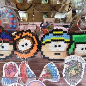 May include: Pixel art keychains in various colors, including blue, orange, and green, depicting cartoon characters. The keychains are displayed alongside colorful bead bracelets and stickers with text and illustrations. The background includes price tags.