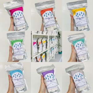 May include: A selection of shower steamers in clear plastic bags, each with a different scent and colour. The bags are labelled with the text "Yours Truly Bath Co." and the scent of each steamer. The steamers are displayed on a shelf.