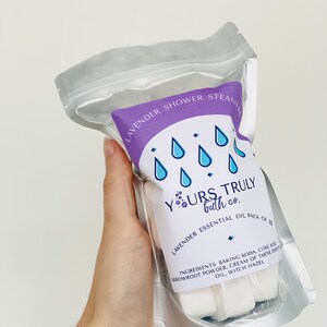 May include: A silver resealable pouch containing 10 white lavender shower steamers. The pouch has a purple label with the text "Lavender Shower Steamers" and "Yours Truly bath co." The label also includes a graphic of four blue raindrops and the text "Lavender Essential Oil Pack of 10". The ingredients are listed on the label: "Baking Soda, Citric Acid, Arrowroot Powder, Cream of Tartar, Essential Oil, Witch Hazel."