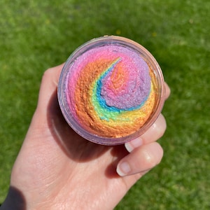 May include: A small jar of rainbow-colored bath scrub. The scrub is a swirl of pink, orange, yellow, green, blue, and purple.