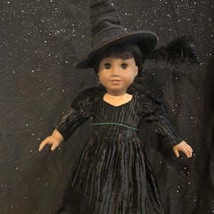 May include: A doll dressed in a black witch costume, including a long-sleeved dress, a pointed hat, and black shoes. The dress has a green accent at the waist. A black feather is attached to the hat. The doll is set against a starry background.