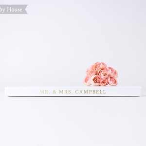 May include: A white book with the text "MR. & MRS. CAMPBELL" on the cover. A bouquet of pink flowers is resting on top of the book.