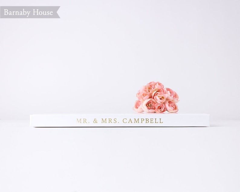 May include: A white book with the text "MR. & MRS. CAMPBELL" on the cover. A bouquet of pink flowers is resting on top of the book.