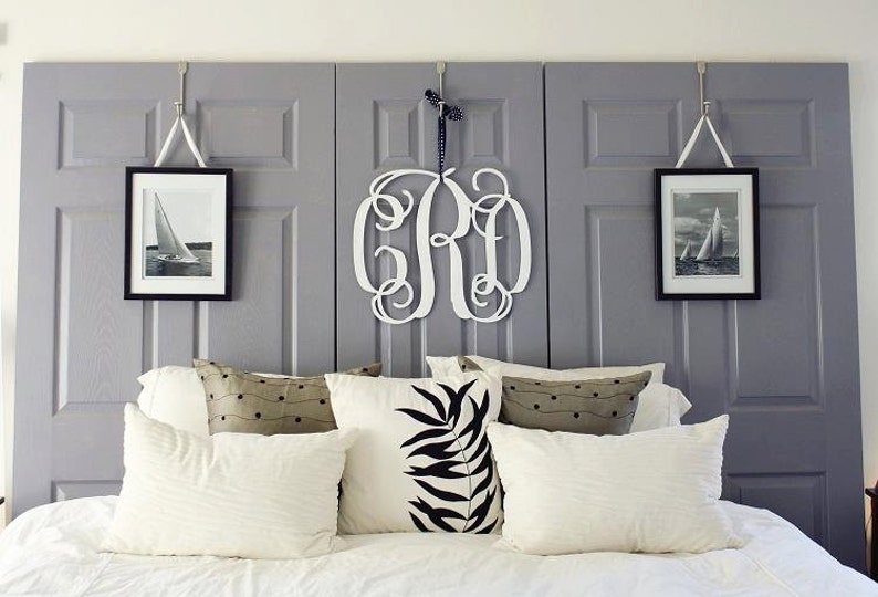 May include: A white bed with a gray headboard made of three repurposed doors. The headboard has two framed pictures of sailboats and a white wooden monogram in the center. The bed has white bedding and several decorative pillows.