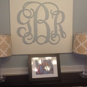 May include: Two beige lamps with white geometric patterned lampshades sit on a dark brown table in front of a white canvas with a large blue metal monogram of the letters "BR".