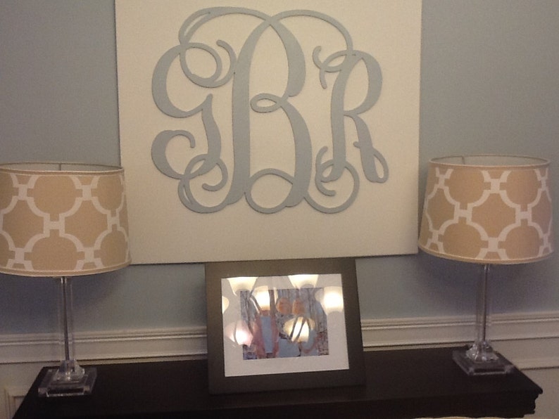 May include: Two beige lamps with white geometric patterned lampshades sit on a dark brown table in front of a white canvas with a large blue metal monogram of the letters "BR".