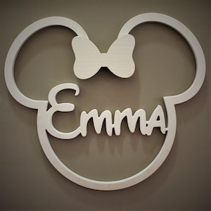 May include: A white wooden wall decoration in the shape of a mouse head with the name "Emma" in cursive script. A bow is centered above the name. The decoration is mounted on a neutral-colored wall.