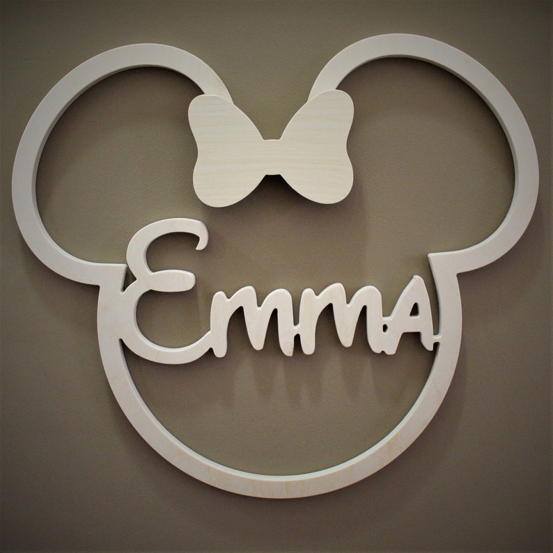 May include: A white wooden wall decoration in the shape of a mouse head with the name "Emma" in cursive script. A bow is centered above the name. The decoration is mounted on a neutral-colored wall.