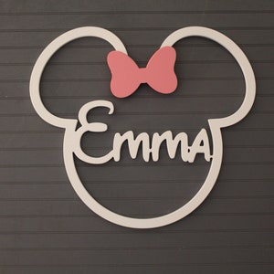 May include: A white wooden wall decoration in the shape of a mouse head with the name "Emma" in cursive script. A pink bow is above the name. The decoration is mounted on a dark gray, horizontally-paneled wall.