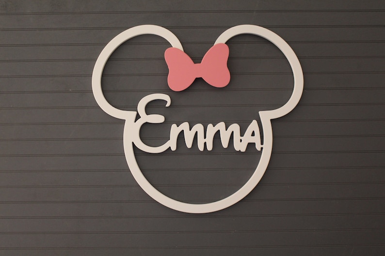 May include: A white wooden wall decoration in the shape of a mouse head with the name "Emma" in cursive script. A pink bow is above the name. The decoration is mounted on a dark gray, horizontally-paneled wall.