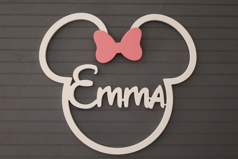 May include: A white wooden wall decoration in the shape of Mickey Mouse ears, with a pink bow and the name "Emma" in cursive. The decoration is mounted on a dark gray, paneled wall.