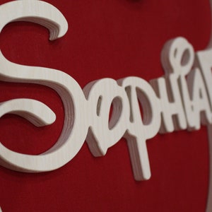 May include: A wooden sign with the name "Sophia" written in a decorative script. The sign is painted white and is on a red background.
