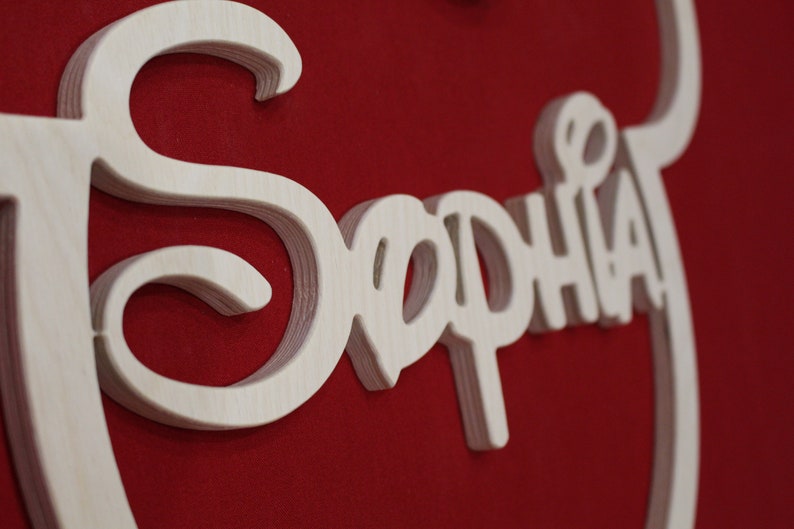 May include: A wooden sign with the name "Sophia" written in a decorative script. The sign is painted white and is on a red background.