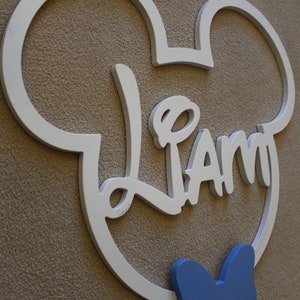 May include: White wooden Mickey Mouse head cutout with the name "Liam" in cursive lettering and a blue bow tie.