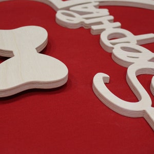 May include: A wooden cutout of Mickey Mouse ears with a bow on the left side. The ears are shaped like a circle with a smaller circle inside. The bow is a simple shape with two loops. The cutout is painted white and is on a red background.