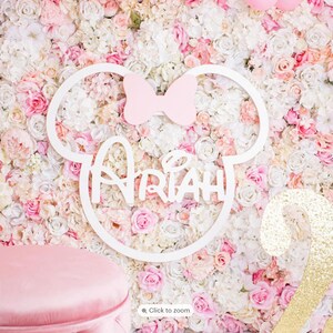 May include: A white wooden Mickey Mouse head cutout with a pink bow and the name "Ariah" written in a whimsical font. The cutout is placed on a wall covered in pink and white artificial flowers.