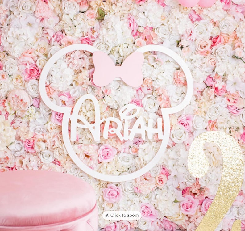 May include: A white wooden Mickey Mouse head cutout with a pink bow and the name "Ariah" written in a whimsical font. The cutout is placed on a wall covered in pink and white artificial flowers.