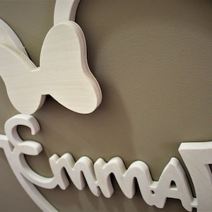 May include: Wooden wall art featuring a Minnie Mouse silhouette with a bow and the name "Emma" in a playful font. The piece is made of light-colored wood and mounted on a neutral-toned wall.