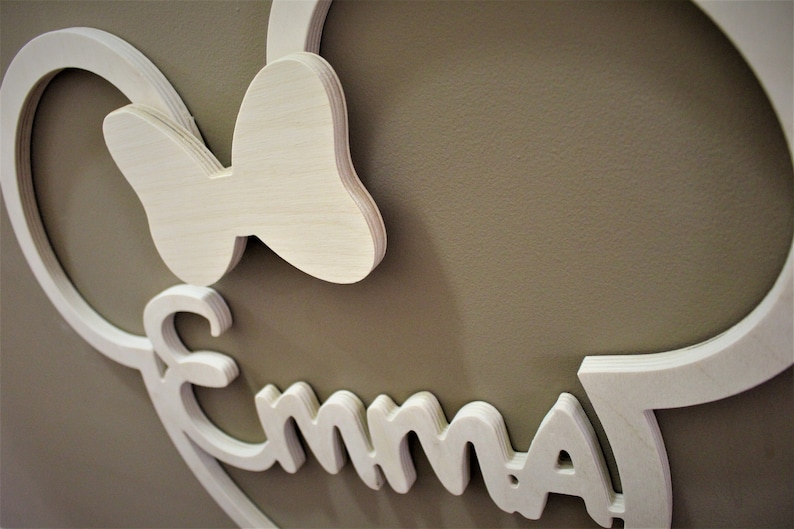 May include: Wooden wall art featuring a Minnie Mouse silhouette with a bow and the name "Emma" in a playful font. The piece is made of light-colored wood and mounted on a neutral-toned wall.