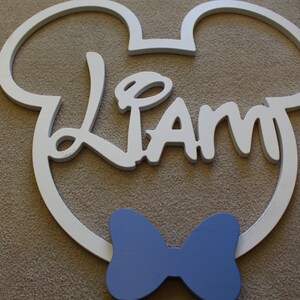 May include: White wooden Mickey Mouse head cutout with the name "Liam" in white script lettering and a blue bow tie.