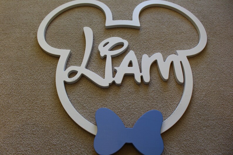 May include: White wooden Mickey Mouse head cutout with the name "Liam" in white script lettering and a blue bow tie.