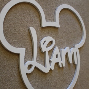 May include: White wooden Mickey Mouse-shaped sign with the name "Liam" written in a whimsical font.