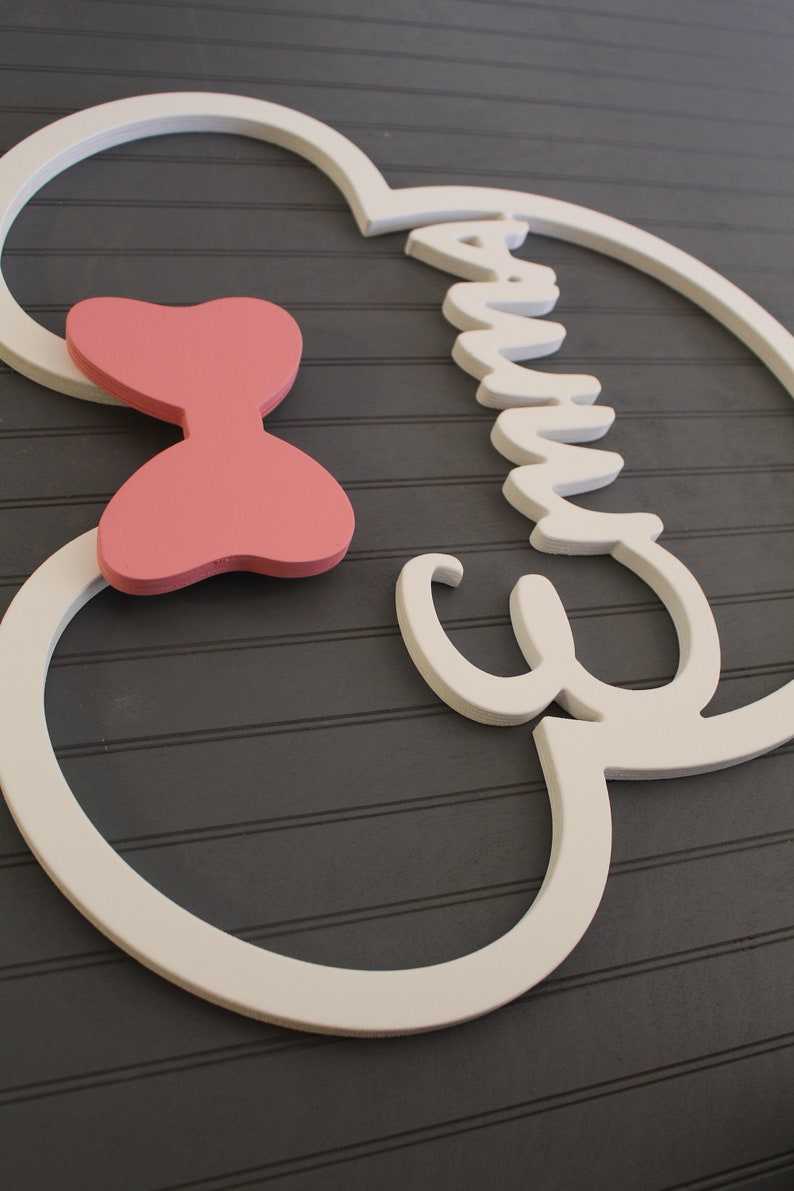 May include: A wooden wall decoration featuring the name "Emma" in white, with a pink bow accent. The design is set against a gray background with horizontal lines, creating a charming and personalized piece.