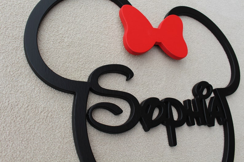 May include: Black wooden cutout of a Mickey Mouse head with the name "Sophia" written in a decorative font. A red bow is attached to the top of the head.