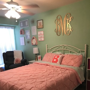 May include: A bedroom with a light green wall, a white bed frame, and a pink comforter. The wall is decorated with a gold monogram, a bear picture, and other wall decor. There is a black armchair with a white throw blanket and a pink flower on it.