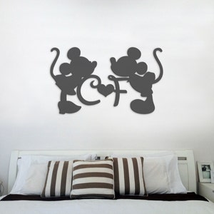 Puede incluir: Decoraci&oacute;n de pared en silueta negra de Mickey Mouse y Minnie Mouse tom&aacute;ndose de la mano con un coraz&oacute;n en el medio. La decoraci&oacute;n est&aacute; montada en una pared blanca sobre una cama blanca con almohadas a rayas marrones y blancas.