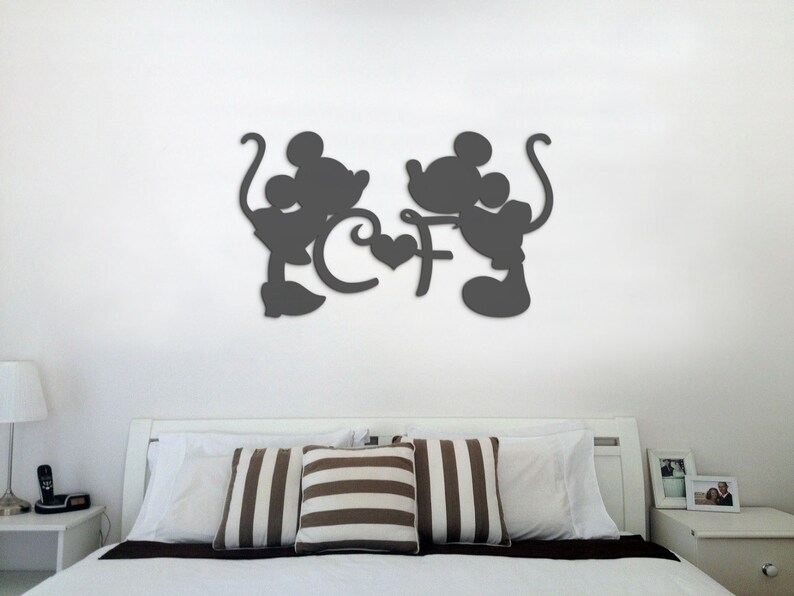 Puede incluir: Decoraci&oacute;n de pared en silueta negra de Mickey Mouse y Minnie Mouse tom&aacute;ndose de la mano con un coraz&oacute;n en el medio. La decoraci&oacute;n est&aacute; montada en una pared blanca sobre una cama blanca con almohadas a rayas marrones y blancas.